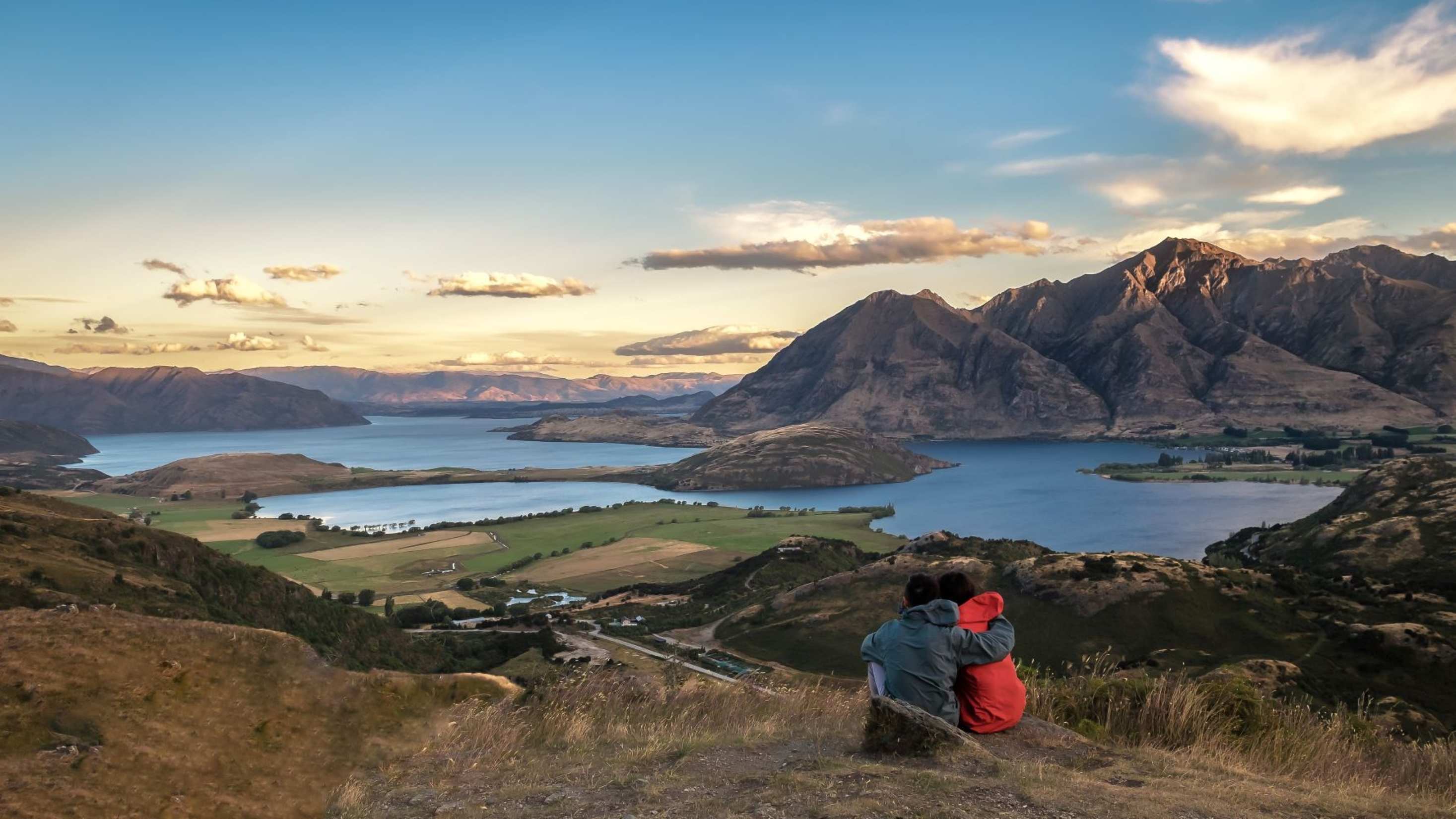 Couple admiring a scenic view of New Zealand's lakes, mountains, and rolling landscapes at sunset.