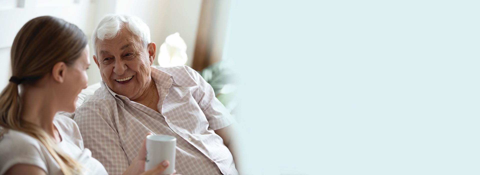 Senior man and young woman sitting together, having a conversation over coffee in a well-lit home setting.