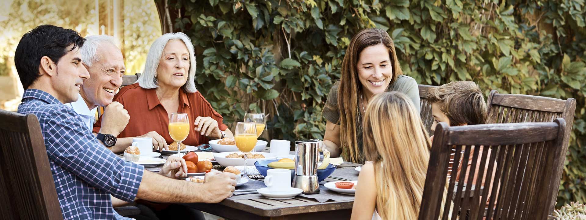 Family enjoying breakfast together outdoors with orange juice, pastries, and fruits on a wooden patio table surrounded by greenery.