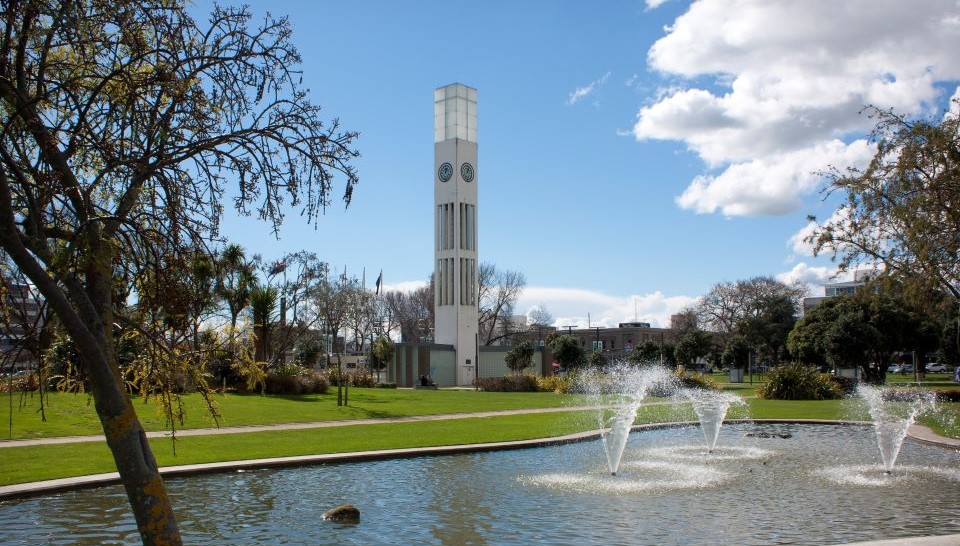 Clock tower in Manawatu town square with a pond and fountains in the foreground on a sunny day.