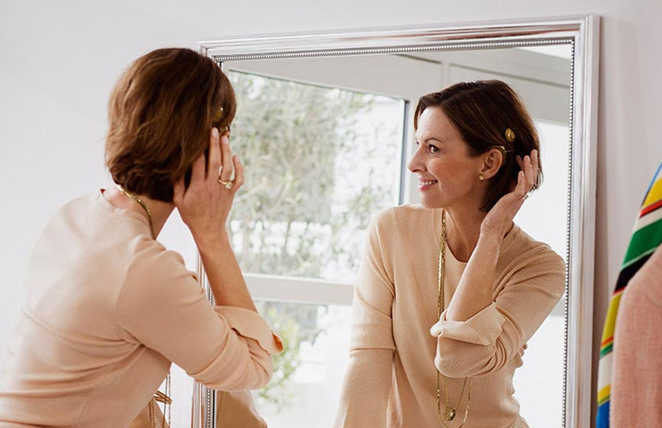 A woman adjusts a discreet beige hearing aid in her ear while looking into a large mirror. She is in a brightly lit room with modern decor and natural light.