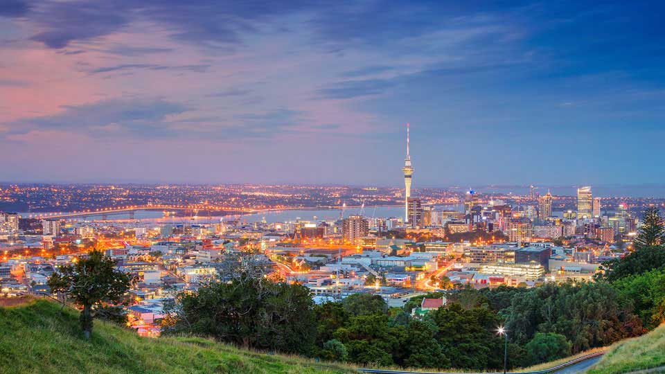 Auckland city skyline at twilight, featuring the Sky Tower, illuminated buildings, and surrounding greenery.