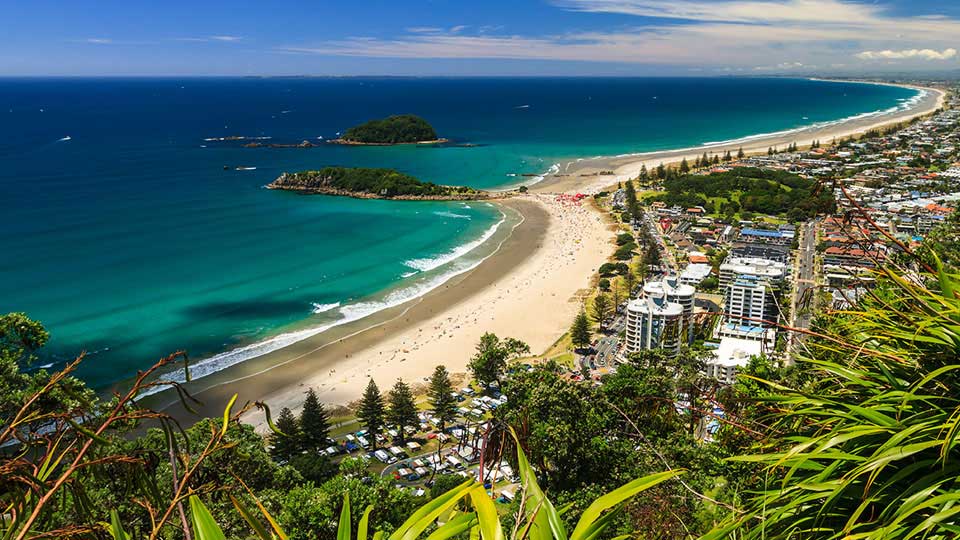 Aerial view of Mount Maunganui beach and coastline in Bay of Plenty, New Zealand, with turquoise water, sandy shores, and nearby urban development.