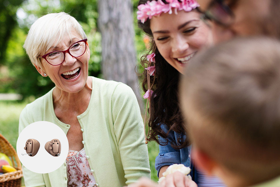 A senior woman, wearing a light green cardigan, interacts with family outdoors in a park setting. Inset shows a pair of discreet in-ear hearing aids, emphasizing effective hearing solutions.