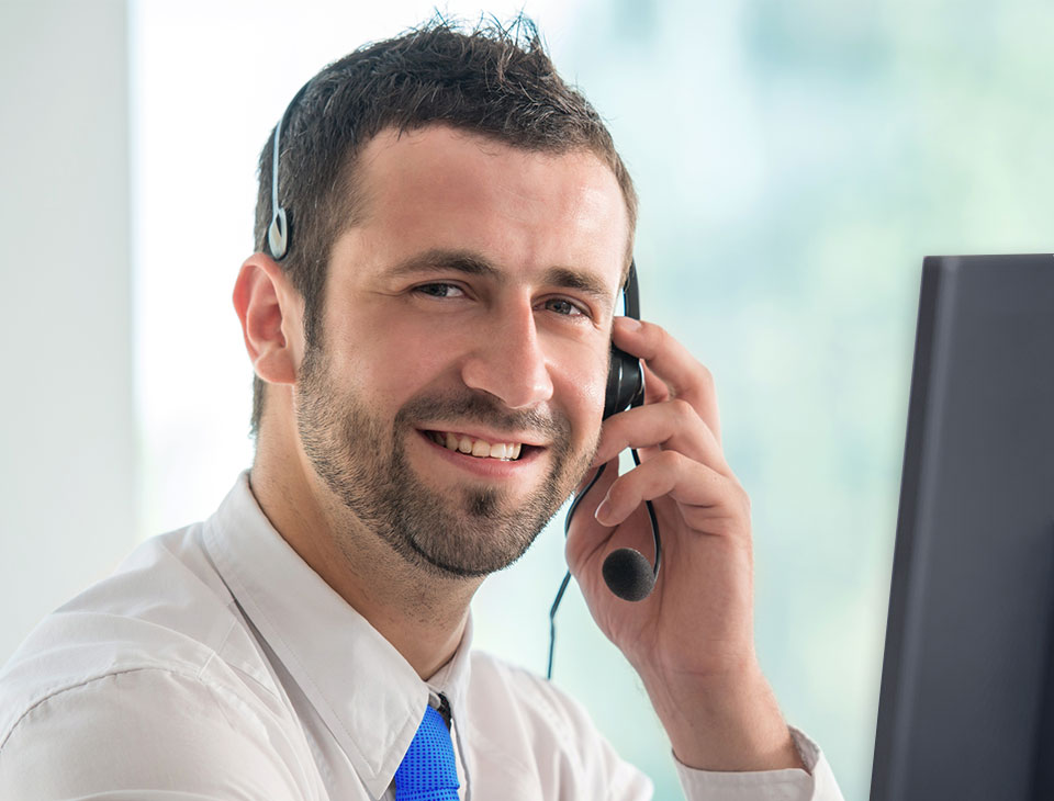 A customer service representative wearing a headset speaks into a microphone, seated in front of a computer within a brightly lit office setting. No visible text or hearing aid branding is present.