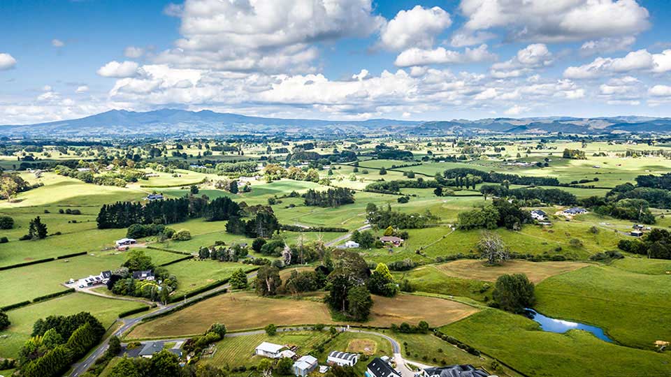 Lush green rural landscape of Waikato, New Zealand, featuring rolling farmland, scattered houses, and distant mountains under a partly cloudy sky.