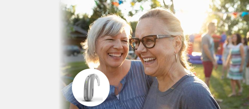 Two older adults embracing outdoors at a sunny gathering, with a hearing aid device displayed in the foreground.