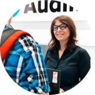 A professional audiologist in a black shirt interacts with a customer wearing a blue plaid shirt and red backpack at an Audika hearing clinic reception desk. The Audika logo is partially visible.