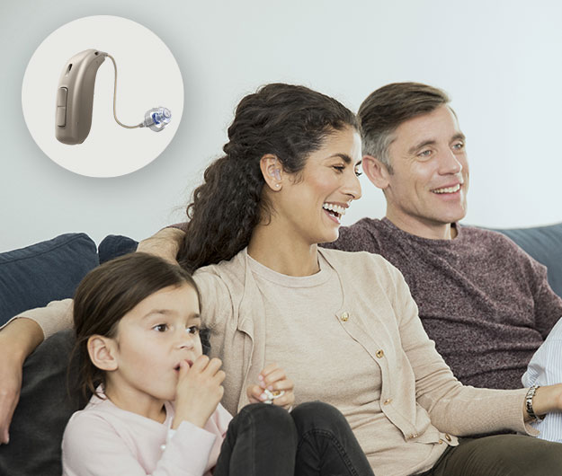 A woman wearing an Oticon hearing aid sits on a couch with her family, enjoying quality time in a cozy home setting. Inset shows a close-up of the hearing aid device.