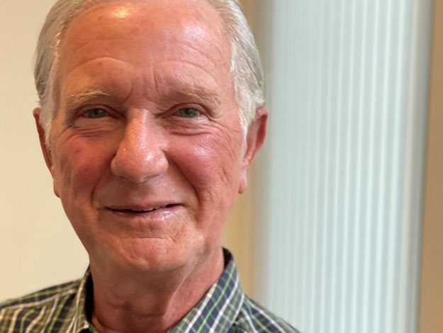 A senior individual with gray hair wears a plaid shirt, standing indoors next to a window with vertical blinds. The image is associated with Audika, a hearing care clinic.