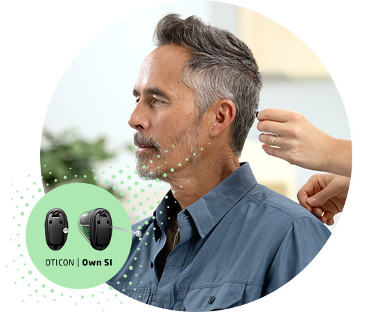 A person with short, salt-and-pepper hair sits in a hearing clinic, as an audiologist adjusts a discreet behind-the-ear Oticon Own SI hearing aid. A hearing aid image is displayed.
