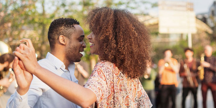 A couple dances outdoors, holding hands, surrounded by blurred musicians playing instruments in a lively park setting. Trees and soft sunlight fill the background, evoking an energetic, communal atmosphere.