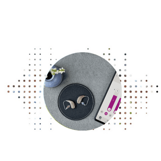 Two beige Oticon hearing aids rest on a leather charging pad beside a smartphone displaying hearing aid controls, set on a minimalist gray surface with a small vase nearby.