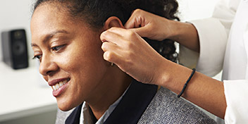 A hearing care professional fits a discreet hearing aid behind the ear of a seated individual in a modern hearing clinic, with a desk and speaker visible in the background.