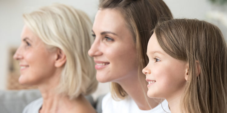 Three generations of women, spanning a grandmother, mother, and daughter, sit closely side-by-side in a well-lit, residential setting, symbolizing family bonding and potentially addressing hearing care solutions.