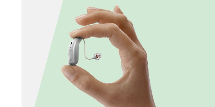 A hand delicately holds a sleek, silver behind-the-ear hearing aid with a thin wire and earpiece, set against a soft, light-green background, illustrating advanced hearing care technology.