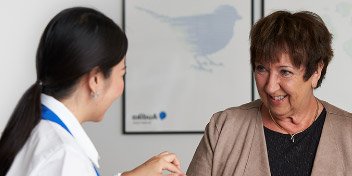 A hearing care professional holds a hearing aid while discussing it with a client in a hearing clinic. The Audika logo and a bird illustration are visible in the background.