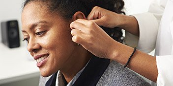 A hearing care professional fits a discreet hearing aid onto a person’s ear in a clean, well-lit hearing clinic. A speaker is visible on a desk in the background.