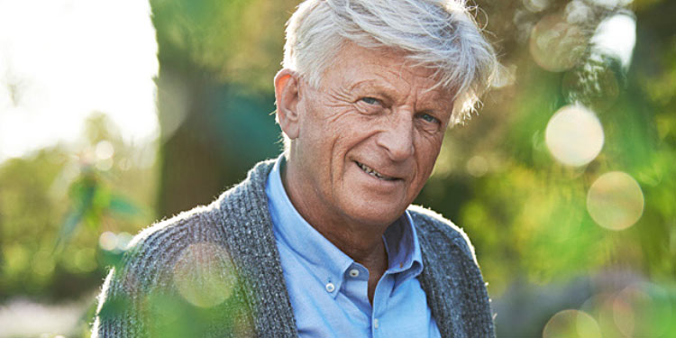 A gray-haired individual wearing a blue shirt and gray cardigan stands outdoors surrounded by greenery, with sunlight creating vibrant lens flares in a natural, serene environment.
