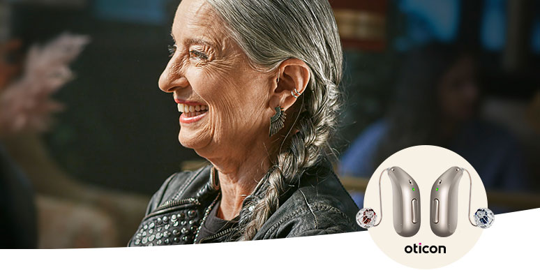 An older woman with braided gray hair wears an Oticon hearing aid discreetly behind her ear, seated in a cozy environment. The image highlights Oticon hearing aids with their sleek, modern design in the foreground, emphasizing advanced hearing care technology.