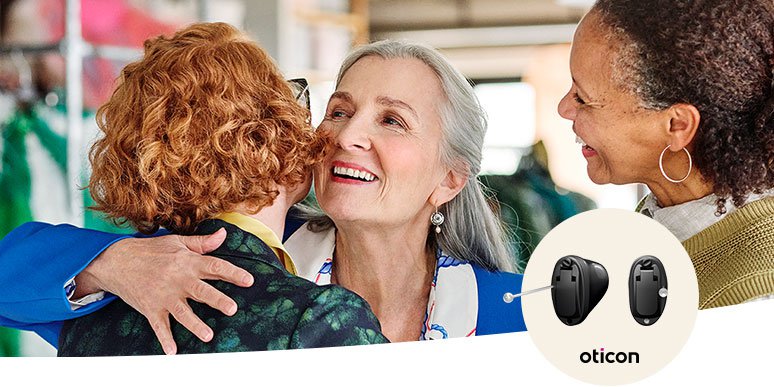 Two individuals embrace warmly in a social setting with blurred faces, while a nearby woman observes. Overlay shows a pair of Oticon hearing aids, highlighting advanced hearing solutions from Audika.