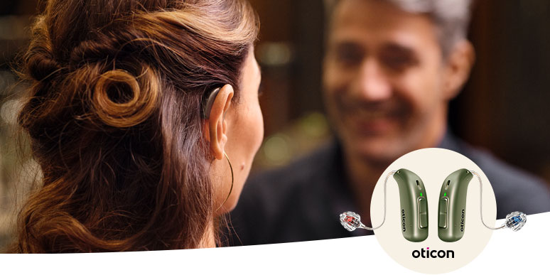 A woman with styled hair converses with another person (face blurred) in a professional setting. Featured inset shows two green Oticon hearing aids with miniRITE design for advanced hearing care.