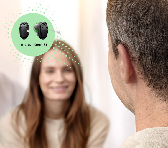 A man wearing a discreet Oticon Own hearing aid listens attentively to a smiling woman during a consultation, with text reading “OTICON | Own S1,” suggesting a hearing clinic setting.