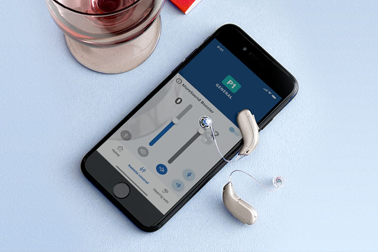 Smartphone displaying a hearing aid control app alongside two modern hearing aids placed on a light blue surface'