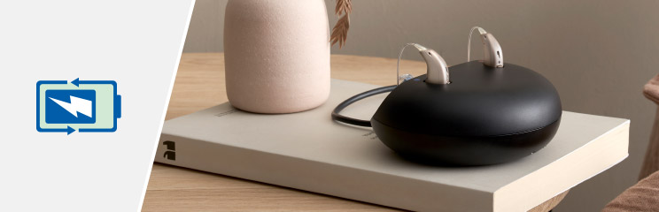 Hearing aid charger with two rechargeable hearing aids placed on a modern table setting, next to a battery charging symbol.