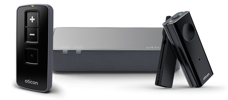 Oticon hearing aid accessories include a sleek black remote control with volume buttons (+/-), a compact gray wireless transmitter, and a clip-on black microphone, enhancing hearing convenience in various settings.