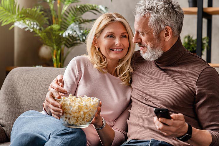 A couple sits closely on a cozy sofa; the woman holds a bowl of popcorn while the man holds a remote control, surrounded by green plants and warm indoor decor.