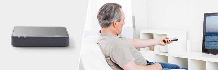 Man sitting on a couch using a TV adapter to enhance hearing device connectivity while watching television.