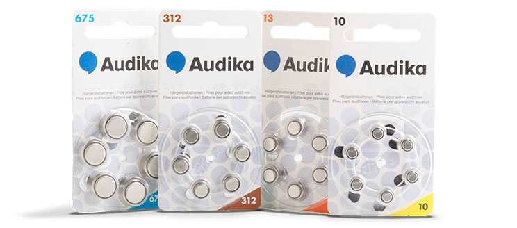 Four packs of Audika-branded hearing aid batteries are displayed, labeled as sizes 675 (blue), 312 (brown), 13 (orange), and 10 (yellow). Each pack contains circular silver batteries arranged in a wheel-shaped plastic holder. The packaging includes the Audika logo featuring a blue speech bubble icon and multilingual text for international accessibility.