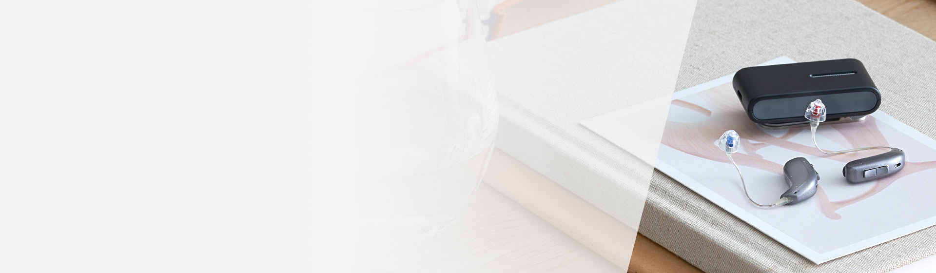 A pair of gray hearing aids with clear earmolds are placed on a modern design booklet next to a black charging case on a beige fabric-covered book in a well-lit setting.