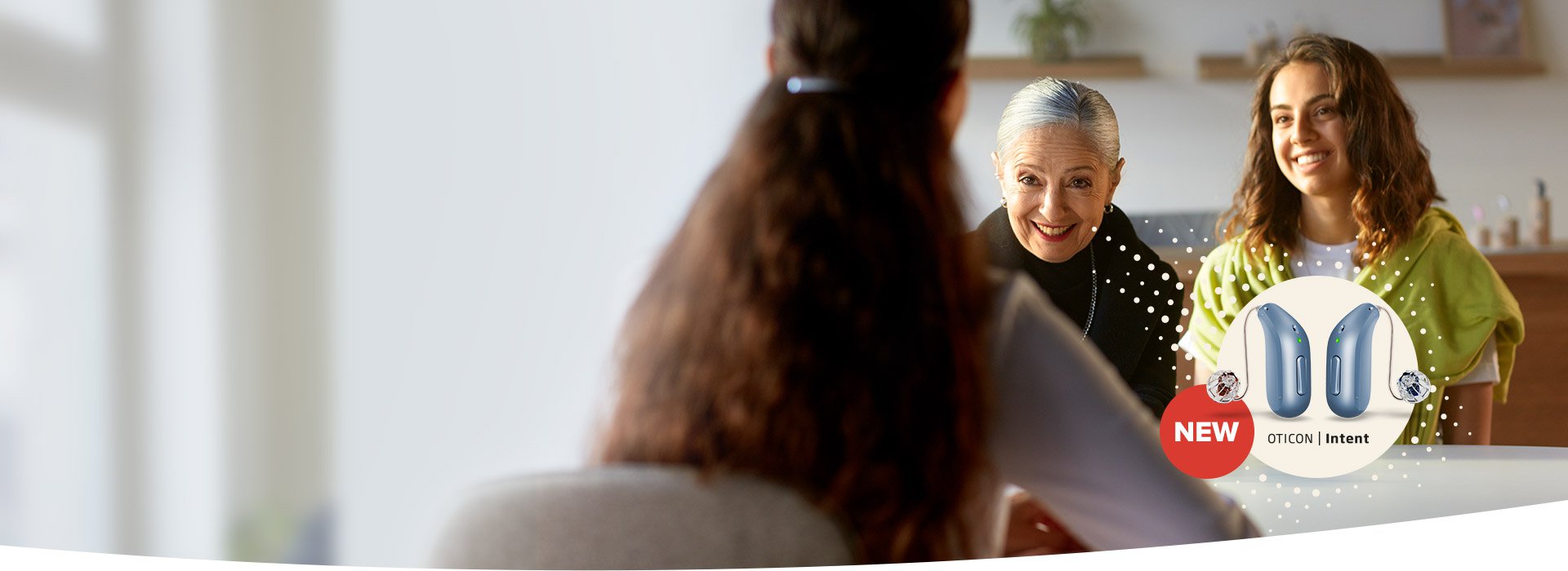 A woman with long, wavy hair sits facing two blurred individuals in a modern clinic setting. The foreground highlights ’NEW OTICON | Intent’ hearing aids, emphasizing hearing care solutions in a welcoming environment.