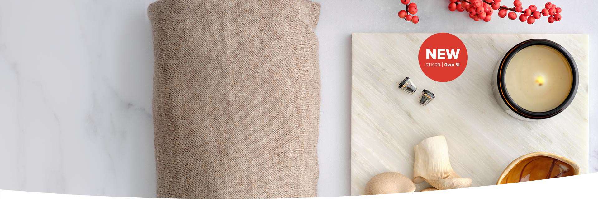 Two Oticon Own 1S hearing aids rest on a marble surface alongside a lit candle, wooden decor, and red berries. A red circle reads: ’NEW OTICON | Own 1S.’