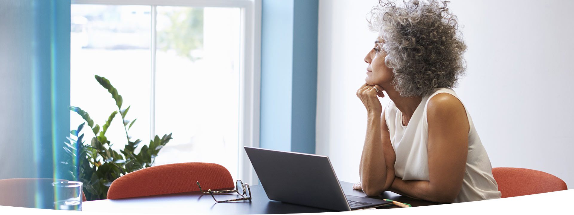 A woman with curly gray hair sits thoughtfully at a desk, resting her chin on her hand, with a laptop, eyeglasses, and glass of water nearby; sunlight filters through a window, and a green plant is visible in the background.
