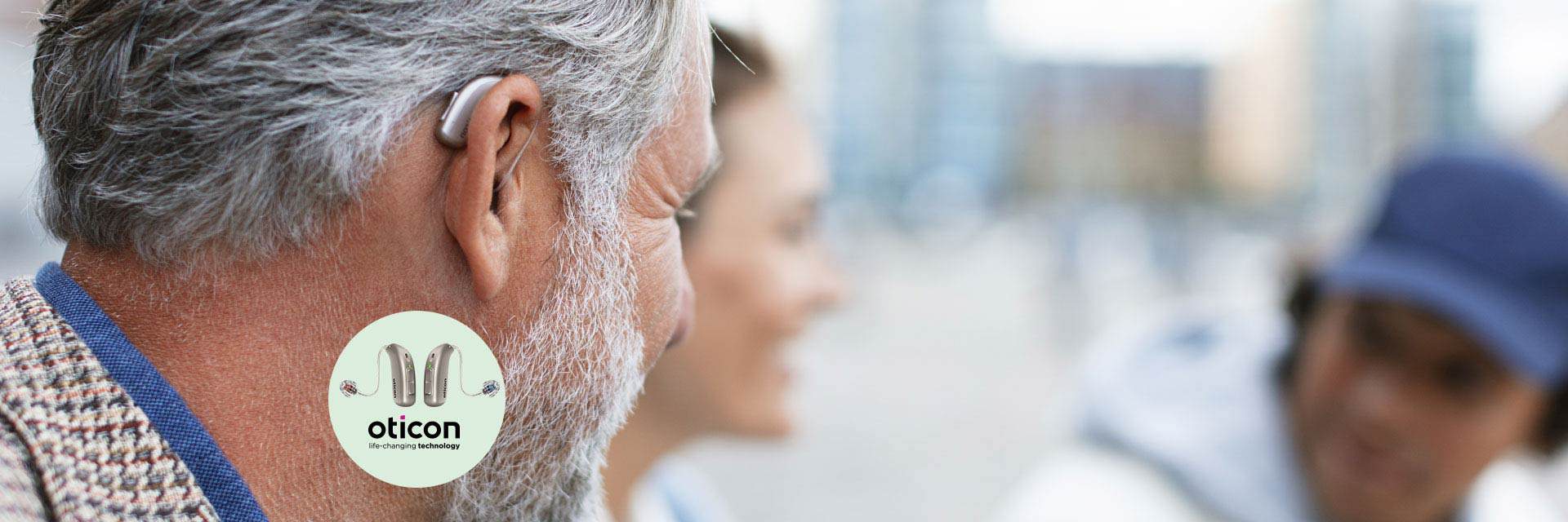 Man with gray hair wearing a discreet hearing aid behind his ear, with the Oticon logo and hearing aid models displayed in the foreground.