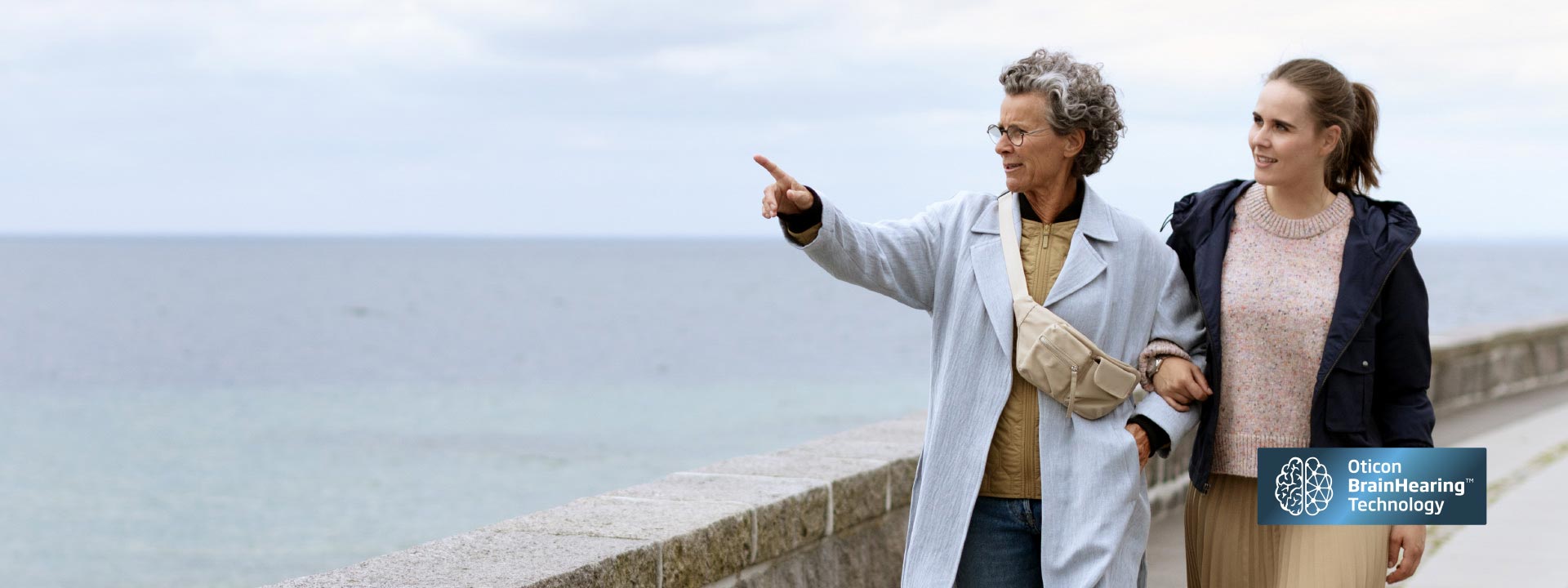 Two women stroll along a seafront walkway, one pointing toward the ocean. The Oticon BrainHearing™ Technology logo appears in the lower-right corner, highlighting advanced hearing aid solutions.