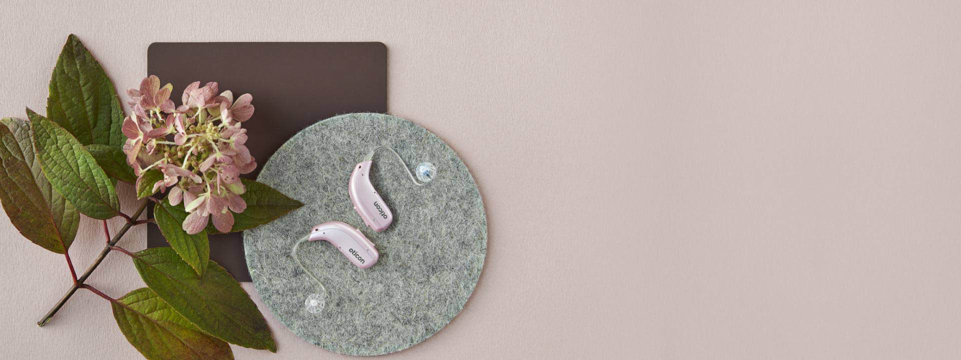 Pink Oticon hearing aids displayed on a textured gray circular mat beside a pale pink hydrangea and green leaves.