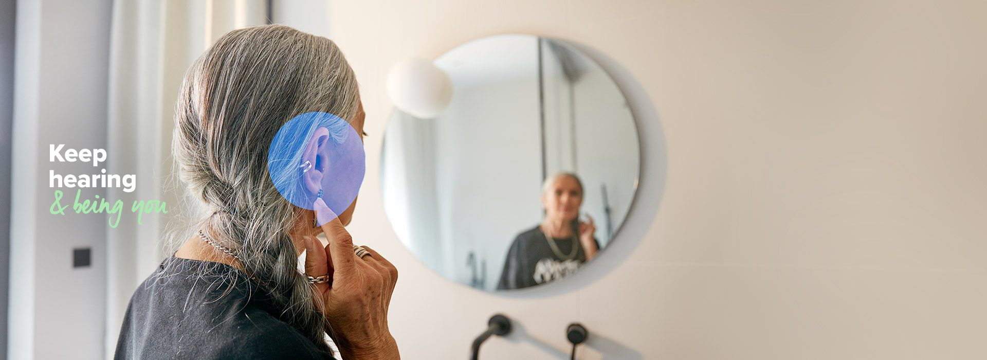 A woman adjusts a discreet hearing aid while standing in front of a circular wall mirror in a modern, softly lit room. Text reads, ’Keep hearing & being you.’