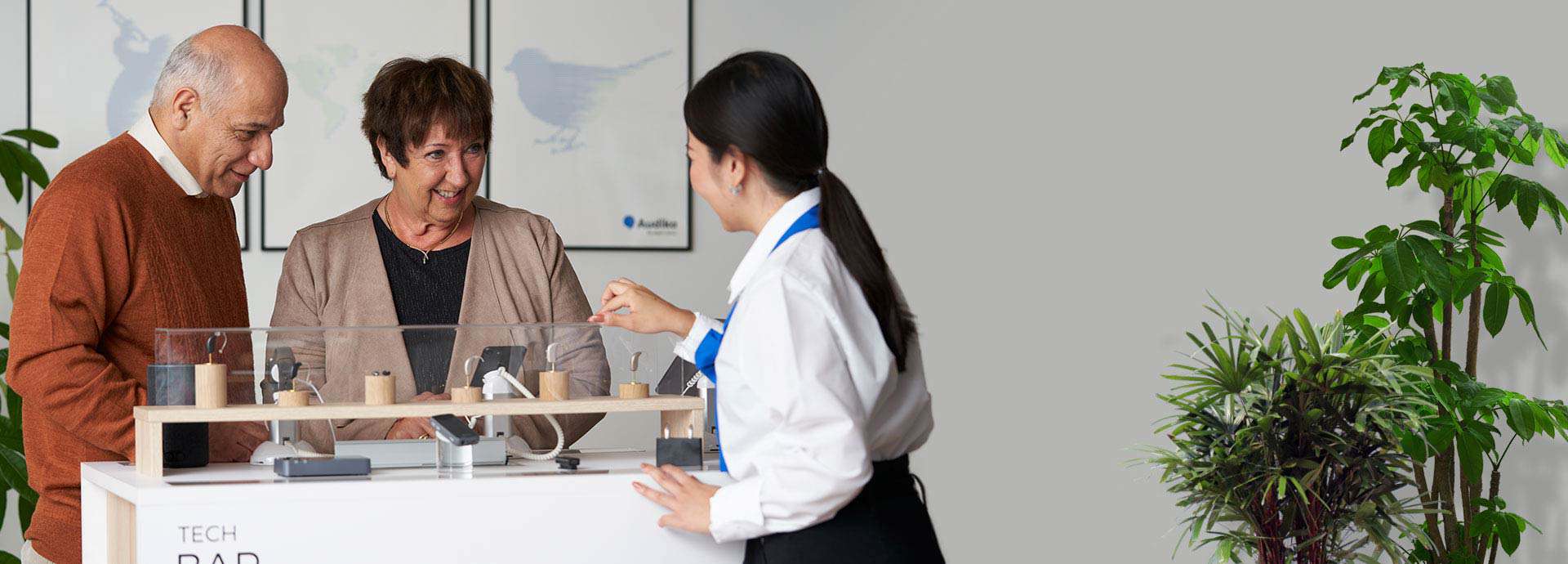 Hearing care specialist demonstrating hearing aid accessories to an older couple at a clinic.