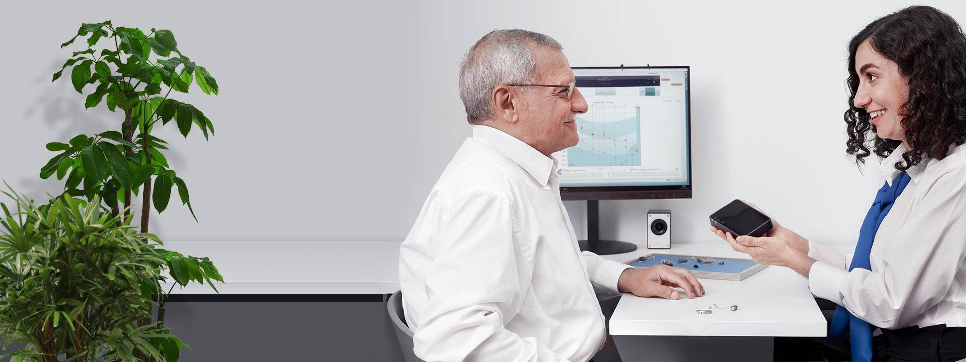 Man consulting with an audiologist in a hearing clinic, sitting near a screen displaying an audiogram and a desk with hearing aids.