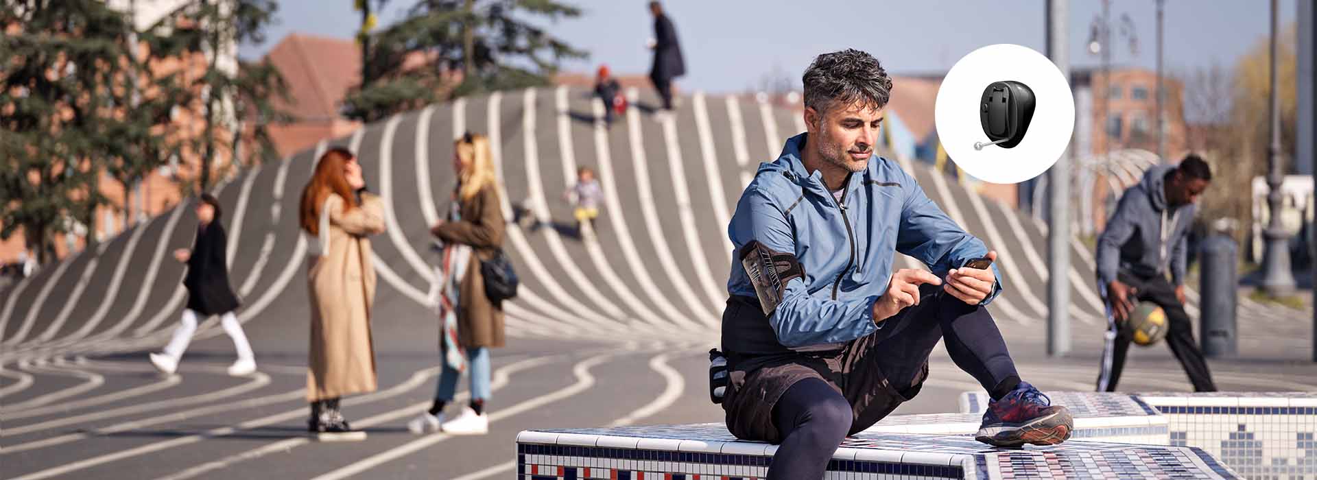 A man sits on a tiled bench in an urban park, holding a phone, wearing a sporty jacket; an Oticon hearing aid icon is overlaid. Others socialize in the background.