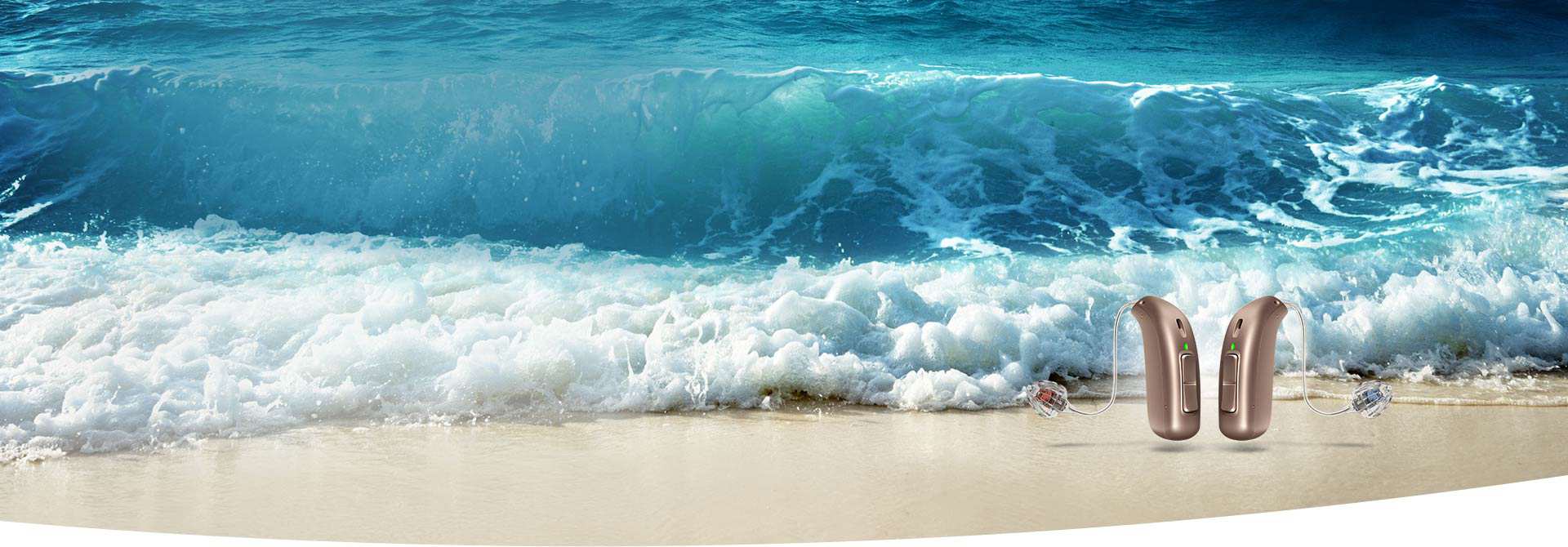 Two sleek, beige hearing aids, likely from Audika, rest on a sandy beach as ocean waves crash in the background, emphasizing clarity and connection in diverse sound environments.