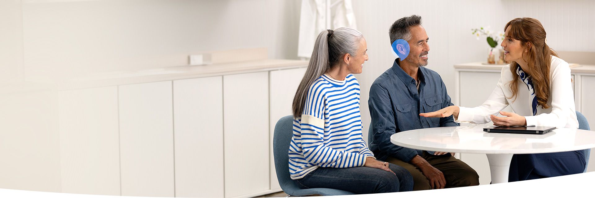 A hearing care professional consults a seated middle-aged couple in a modern hearing clinic. The man wears an in-ear hearing aid, potentially from Oticon or similar brands. A tablet rests on the table.