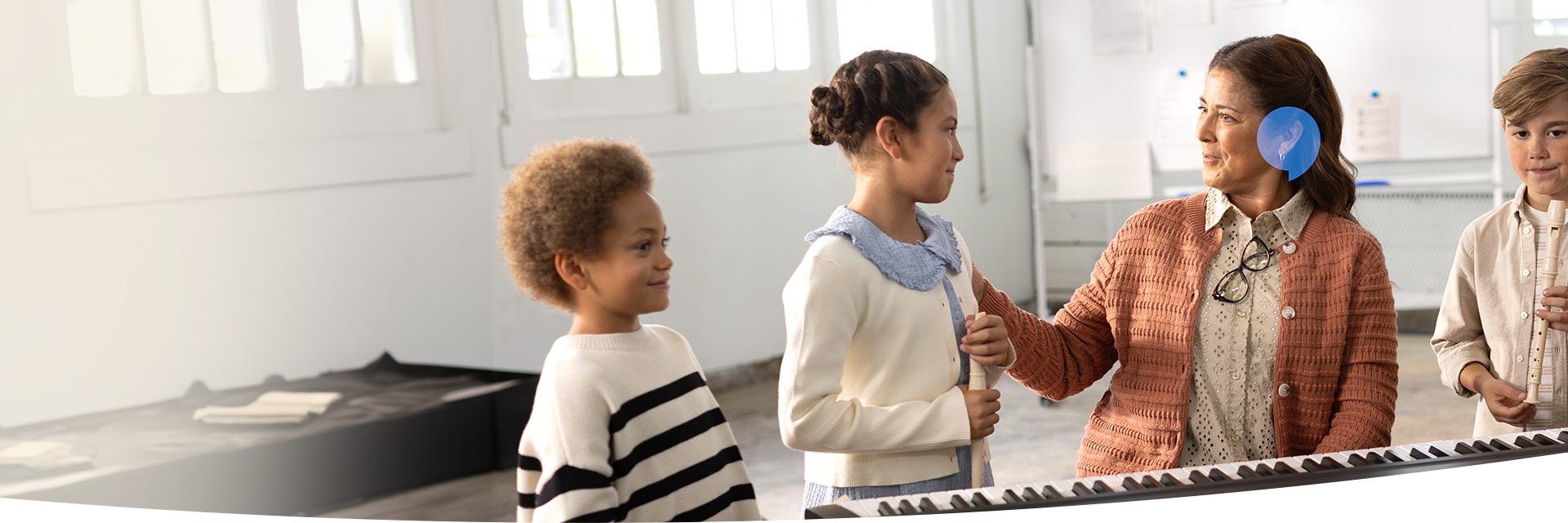 A teacher wearing a hearing aid, possibly from Oticon or Bernafon, is engaging with three children near a keyboard in a bright classroom setting.