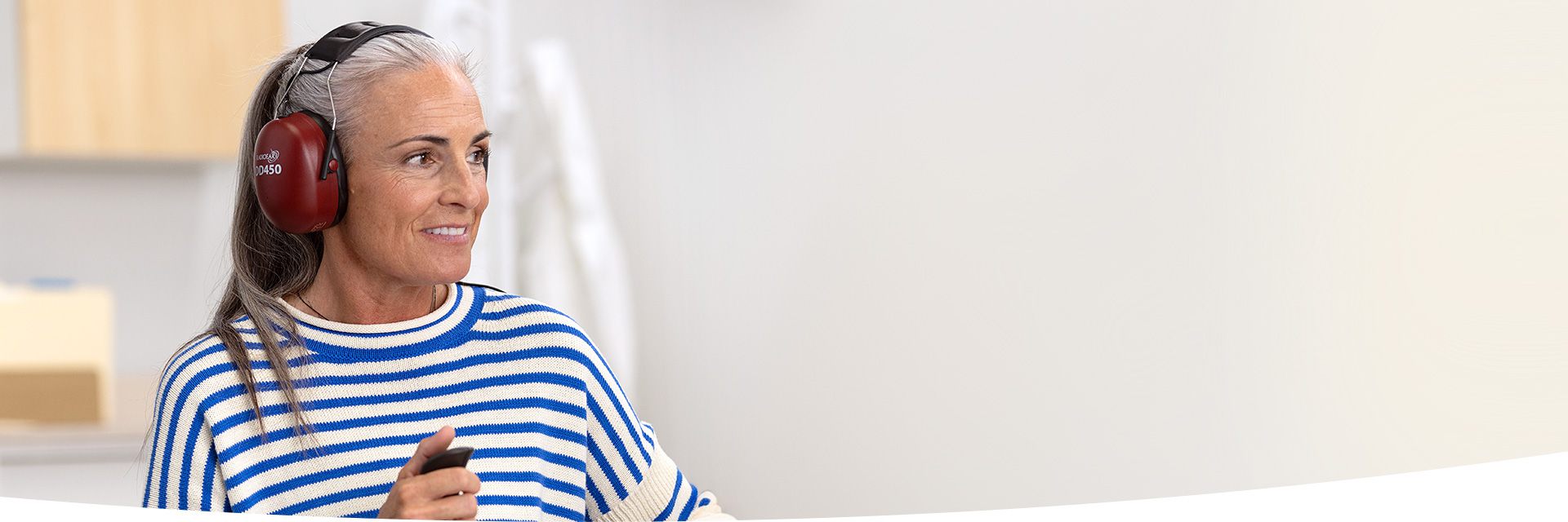 A person wears red ear protection headphones labeled 'Ecklund DD450' while holding a small black device in a bright clinic environment, possibly during a hearing test at a hearing care facility.