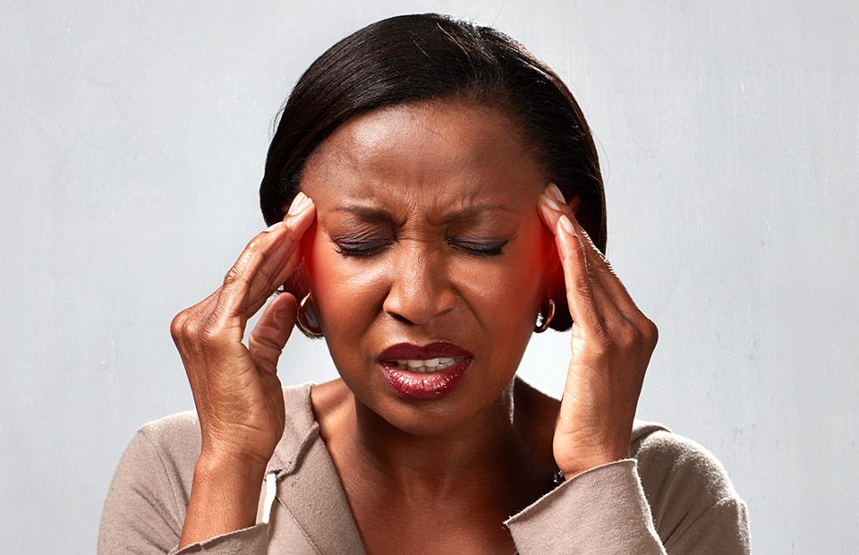 A woman with medium brown skin holds her hands near her ears in a neutral pose, against a plain gray background. The image context suggests hearing care or hearing aid services. No visible text or identifiable hearing aid brands are present.