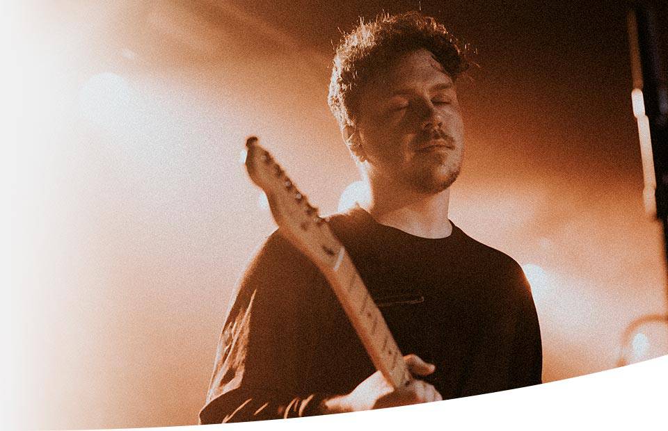 A guitarist performs under warm stage lighting, holding an electric guitar, with minimal background detail suggesting a live concert setting. No text or branding is visible in the image.
